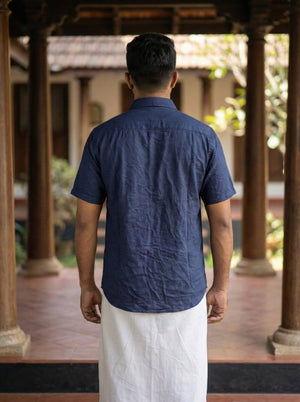 A person stands facing away in a courtyard with wooden pillars and tiled flooring, wearing the Utopian Shirt—a blue short-sleeved bohemian top—and a white traditional garment.