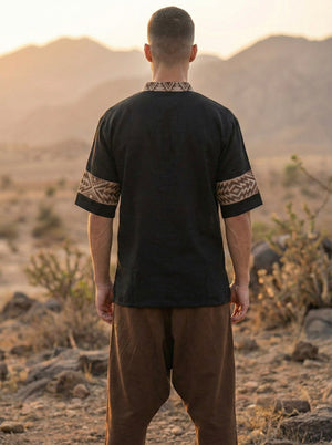 A person stands outdoors facing away, wearing the Aho Kurta—a black shirt featuring traditional Rajasthani handblock print on the cuffs and collar—paired with brown pants. The background showcases dry desert terrain with rocks and distant mountains illuminated by sunset.