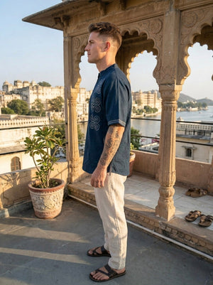 A man wearing the Midnight Nomad kurta, featuring handblock prints in blue, paired with beige pants, stands on a balcony with ornate arches overlooking historic buildings and water on a sunny day, embodying contemporary nomadic fashion.