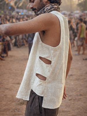 A person wearing the Desert Drift Vest—a sleeveless beige raw weave piece featuring three horizontal side slits—stands outdoors at a crowded event or festival, with blurred people and banners in the background, embodying an effortless festival essential.