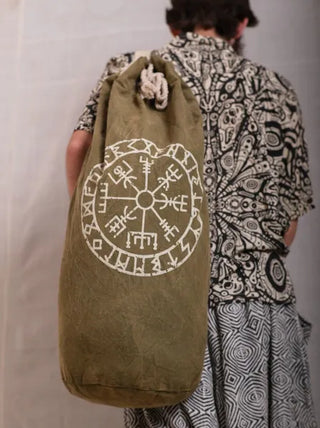 A person stands with their back to the camera, wearing a patterned outfit and carrying a green Vegvisir Duffle bag featuring a white Norse symbol—perfect for secure storage on any travel adventure.