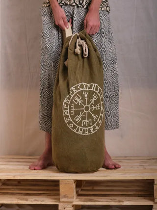 A barefoot person stands on a wooden pallet, holding the Vegvisir Duffle bag—an olive-green bag with white Nordic runes and symbols. This duffle offers secure storage, making it the perfect travel companion for any adventure.