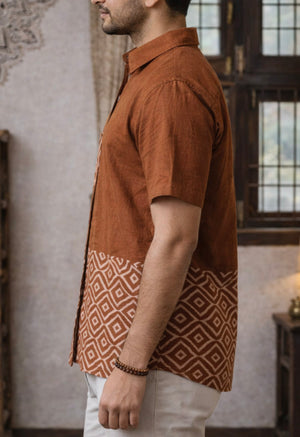 A man stands sideways indoors near a window, wearing the Uday Shirt—an earthy rust hand block printed shirt featuring a geometric block print pattern on the lower half—paired with light-colored pants and a beaded bracelet.