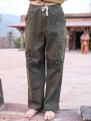 A person stands barefoot outdoors in olive-green 100% cotton Tribal pants, featuring black geometric knee embroidery and a cream drawstring. The background shows a courtyard with another person blurred in the distance.