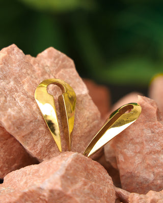 A pair of Chronos Ear Weights in shiny gold teardrop shapes are displayed on rough pink stones, set against a softly blurred green background.