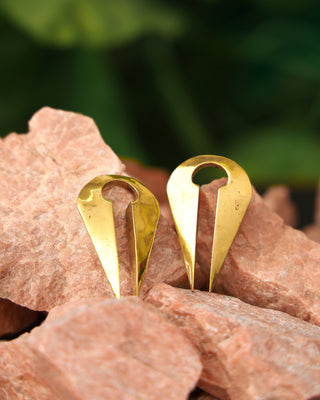 Two shiny, gold-colored Chronos Ear Weights, each teardrop-shaped with a circular cutout at the top, rest on rough light brown stones against a softly blurred green background.