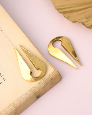 Two gold Chronos Ear Weights, shaped like abstract teardrops, rest on the corner of an open book against a light pink background, with part of a dried leaf visible.