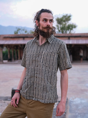 A man with curly hair and a beard is wearing the Tribal Triangle shirt with a Rajasthan-inspired print, paired with khaki pants, as he stands outdoors among trees and rustic buildings.