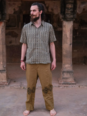 A barefoot man with a beard stands on stone ground in front of an old stone structure with arches, wearing the Tribal Triangle shirt—a unisex top featuring a vibrant Rajasthan print—paired with tan pants adorned with black designs.