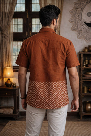 A man stands indoors with his back to the camera, wearing the Uday Shirt—an earthy rust hand block printed shirt adorned with white geometric patterns at the bottom—paired with crisp white pants. The room is decorated with wooden furniture, shelves, and warm lighting.
