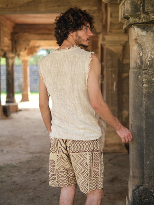A man with curly hair wearing the Shipibo Tank Top—a sleeveless, frayed beige tank with fringe—paired with patterned shorts stands outdoors near ancient stone pillars, looking to the side.