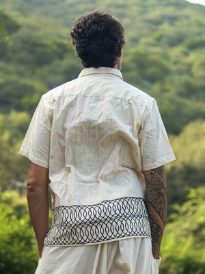 A person with dark, curly hair stands outdoors, viewed from behind, wearing the Vairagya Shirt—a light short-sleeve shirt made from organic cotton and adorned with a dark geometric pattern along the hem; lush green trees and hills fill the background, embodying a dedication to conscious fashion.