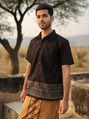 A man stands outdoors in a warm, natural setting with a tree and grass in the background, wearing the Maja Shirt—a black short-sleeve bohemian shirt with geometric patterns—paired with tan pants featuring designs.