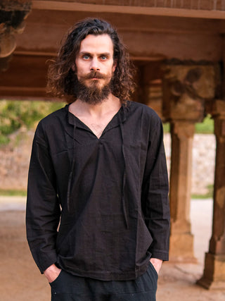 A man with long curly hair and a full beard stands outdoors under a wooden structure, hands in his pockets, wearing the brittany naim kurta—a loose-fitting black shirt—paired with size 46 black pants (30 length), looking at the camera.