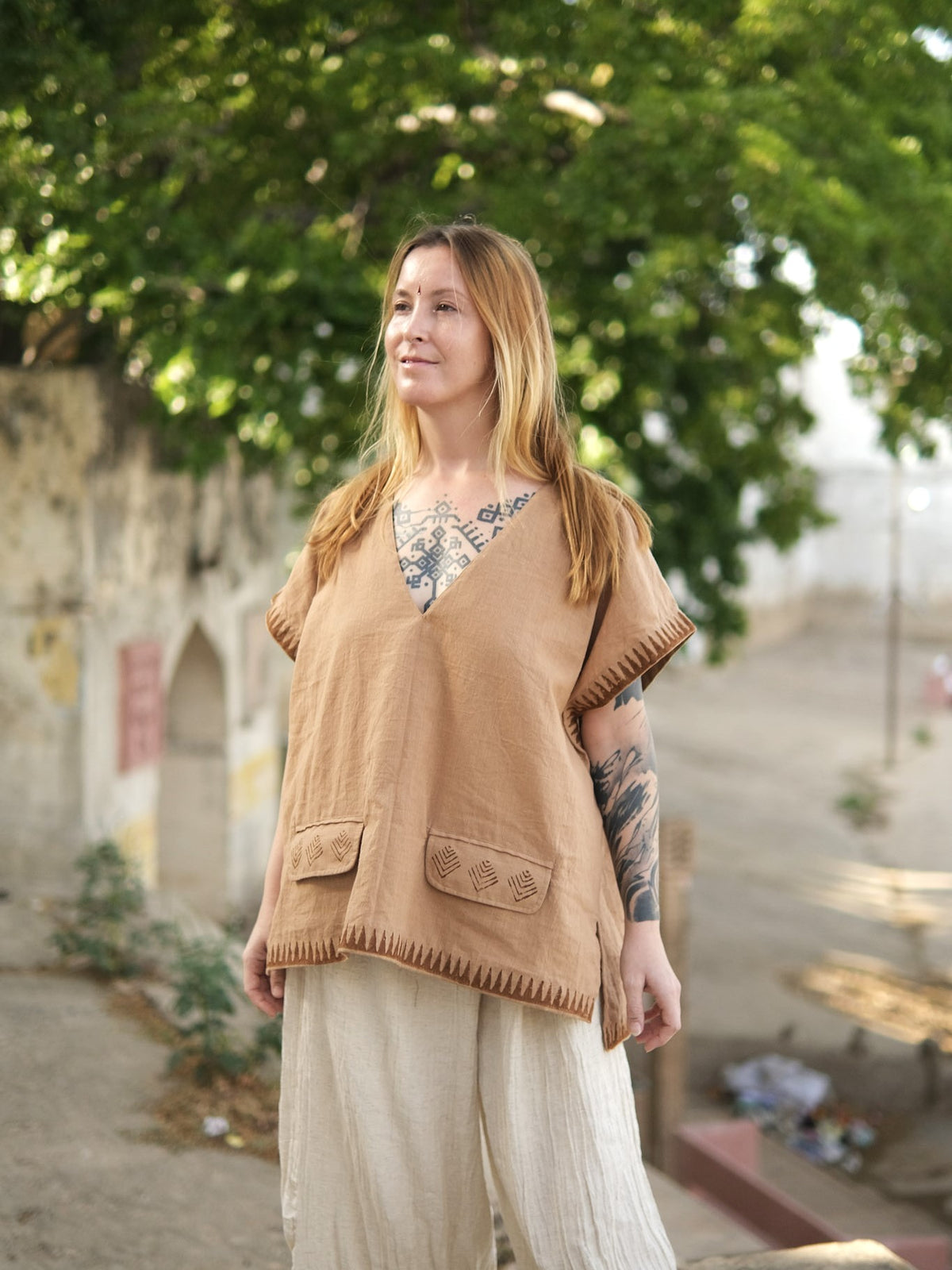 A woman wearing the tan, v-neck Mysa Vest in a boho style with embroidered pocket flaps and a geometric hemline.