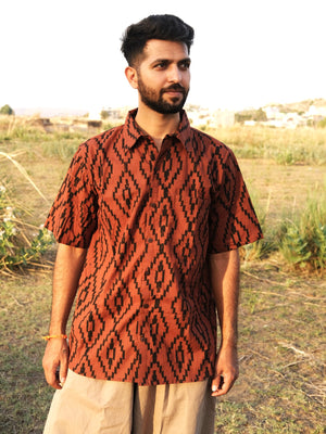A man stands outdoors looking to the side, wearing the Terraza Shirt—a tribal-inspired geometric short-sleeve shirt in brown and black—paired with beige pants, with grass and a blurred landscape in the background.
