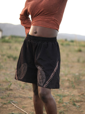 Wearing Kaari Leaf Shorts featuring handcrafted leaf motifs and a long-sleeve rust-colored shirt, the person stands on dry, grassy ground outdoors.