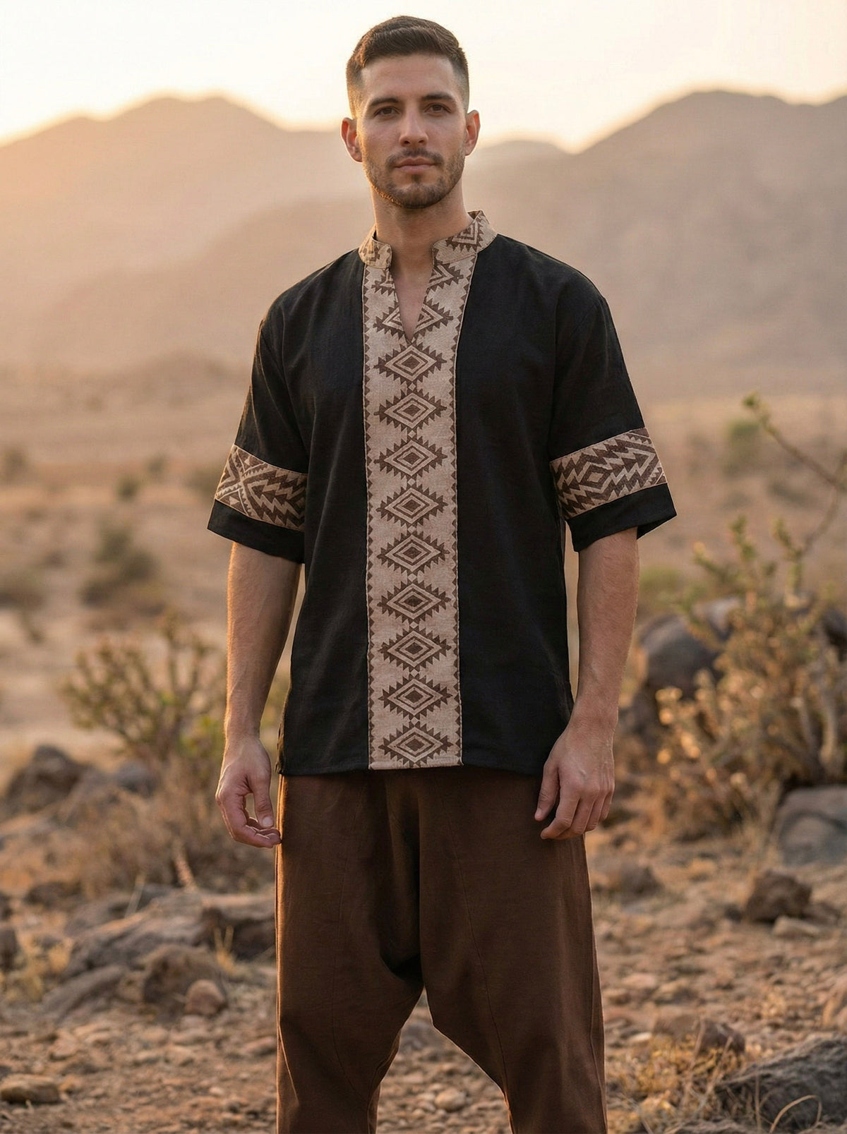 A man stands outdoors at sunset in a desert landscape with rocky hills and plants, wearing a black Aho Kurta featuring beige geometric Rajasthani handblock prints.