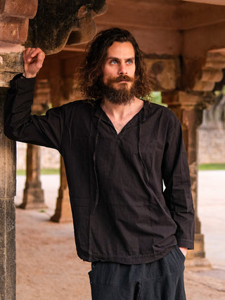 A man with curly brown hair and a beard, wearing the Naim Kurta and matching sustainable cotton pants in black, leans against a stone pillar in an ancient outdoor structure with arches.