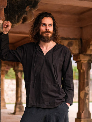 A man with long curly hair and a beard, wearing the Naim Kurta—a loose-fitting piece made from sustainable cotton—stands under stone arches, one arm resting on a column as he gazes into the distance.