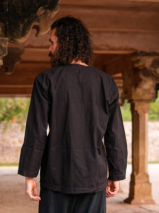 A person with curly hair, seen from behind under a wooden structure, wears the loose-fitting black Naim Kurta and dark pants, set against a backdrop of stone and greenery that highlights the kurtas timeless style.