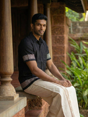 A man wearing the Adhir Shirt—crafted from black cotton with patterned sleeves—pairs it with a cream-colored traditional garment as he sits on a stone ledge outside, surrounded by pillars and green plants, presenting an ethnic contemporary look.