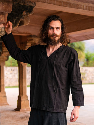 A man with curly hair and a beard wears the loose black Naim Kurta, made from sustainable cotton, standing outdoors beneath a stone structure, with trees and a stone wall behind him.