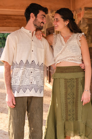 A man and woman stand outdoors, smiling. The man wears a Syan Shirt with a subtle block print and olive pants, while the woman looks stylish in a cream crochet top and long olive skirt. Both appear happy and relaxed together.
