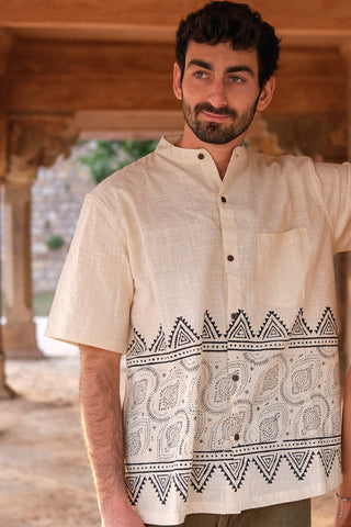 A man with short dark hair and a beard stands outdoors, smiling softly as he leans against a pillar, wearing the stylish Syan Shirt featuring black geometric and paisley block printed designs near the hem.