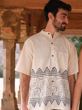 A man with short dark hair and a beard wears the stylish Syan Shirt, a cream short-sleeve top featuring a black block print. He stands outdoors under a stone structure, looking to his right and smiling.