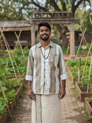 A man wearing the Dwara Shirt, an artisanal block print cotton shirt, and a traditional white dhoti stands smiling in a garden with raised plant beds, lush greenery, trees, and a stone archway in the background.