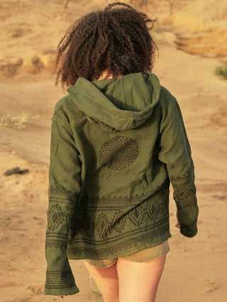 In a sandy desert landscape, a person with curly hair embraces a boho vibe wearing the Tribal Hoodie in olive green, showcasing geometric patterns. Khaki shorts complete their look against the warm, arid backdrop.