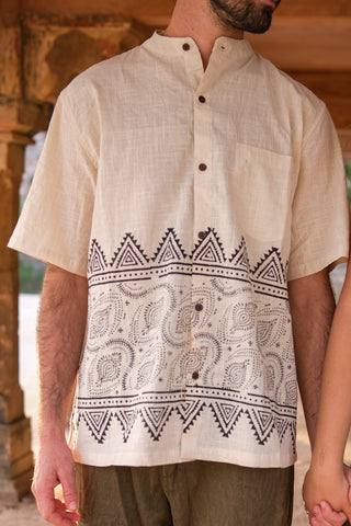 A man wears the stylish Syan Shirt—a cream short-sleeve shirt featuring a mandarin collar, brown buttons, chest pocket, and a black block-printed geometric and paisley border at the hem; only his torso and part of his arm are shown.