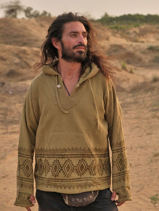 A person with flowing hair and a beard stands in the desert, wearing a sand yellow Tribal Hoodie. They gaze sideways with a calm expression, surrounded by sandy terrain and sparse vegetation that accentuates their boho babe vibe.