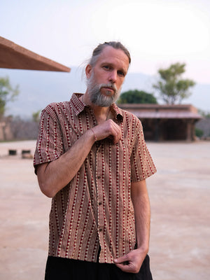 A bearded man with a mustache stands outdoors on a sunny day, holding the chest of his patterned short-sleeve Tribal Triangle shirt. The unique unisex Rajasthan print of the Tribal Triangle shirt adds a distinctive flair to his casual style, while trees and buildings blur in the background.