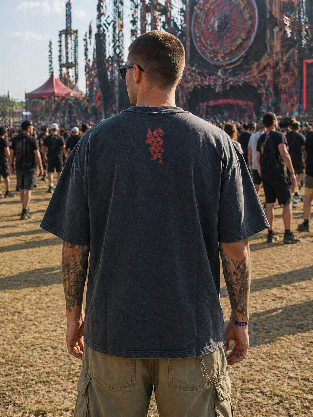 A man with tattoos, wearing the Dragon Oversized Tee and cargo shorts, stands with his back to the camera at an outdoor music festival, surrounded by other attendees near a large decorated stage. The look channels effortless streetwear vibes amidst the lively crowd.