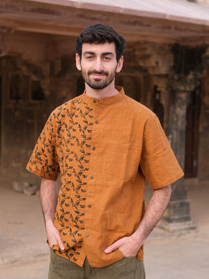 A man with short dark hair and a beard stands outdoors, hands in pockets, wearing the Kunal Shirt—an orange statement shirt with a leafy pattern on one side—against an ancient stone building backdrop.