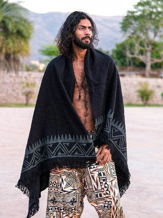 A man with long curly hair and a beard stands outdoors wearing the Oromo Shawl, a black cotton wrap featuring geometric patterns inspired by African tribal art, along with patterned pants. Trees and mountains are visible in the background.