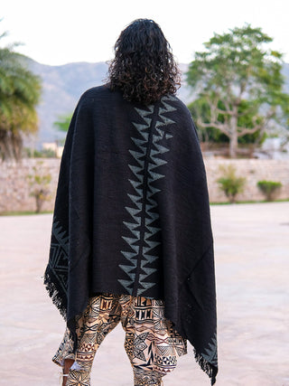 Standing outdoors with curly hair facing away, the person wears the Oromo Shawl—black cotton with a geometric zigzag pattern inspired by African tribal art—and patterned pants, with trees and mountains in the background.
