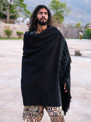 A man with long curly hair and a beard stands outdoors, draped in an Oromo Shawl over his shoulders and wearing geometric-patterned pants. Trees, a stone wall, and distant mountains are in the background.