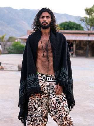 A man with long curly hair and a beard stands outdoors in patterned pants and an Oromo Shawl, adorned with geometric designs inspired by African tribal art, with majestic mountains and trees in the background.