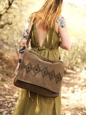 A person in a sleeveless olive green dress with tattoos on their arms walks outdoors, carrying the Safarika Bag—a brown bohemian travel bag with a geometric pattern—over one shoulder.