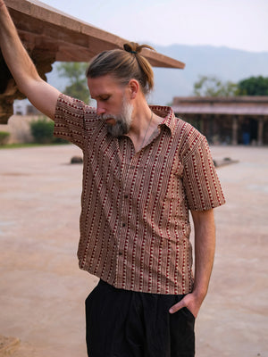 A man with a beard and tied-back hair stands outdoors, leaning against a structure with one hand while looking down. He wears the Tribal Triangle shirt—a unisex piece showcasing a distinct Rajasthan print—paired with dark pants. In the background, distant buildings and trees are visible.
