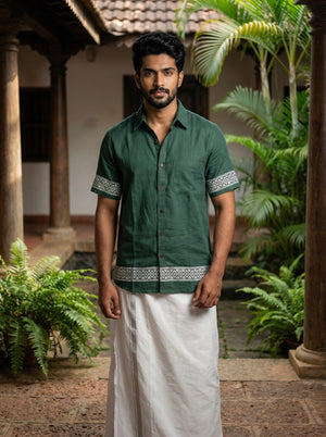 A man wearing the Adhir Shirt in green with patterned borders and a white dhoti stands in a traditional courtyard featuring plants and wooden pillars.