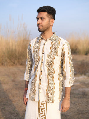 A man stands outdoors in front of tall dry grass, wearing the Dwara Shirt—an artisanal cream and brown top with geometric patterns—paired with a matching dhoti. He looks to the side with a neutral expression beneath a clear sky.