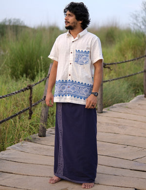 A man stands barefoot on a wooden pathway, dressed in the Bohemian Tribal Shirt—crafted from breathable cotton and adorned with blue block print patterns—and paired with a dark blue traditional wrap skirt. His outfit blends comfort with artisanal style, set against a backdrop of green grass and wooden railings.
