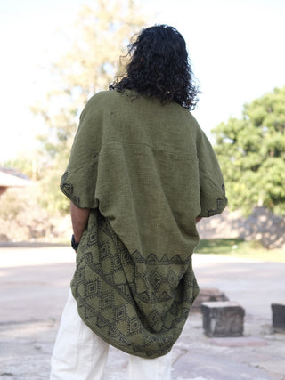 A person with long curly hair wears an olive green Cocoon Shrug with black geometric patterns and light cotton pants, standing outdoors on a sunny day with their back to the camera. Trees and buildings are visible in the background.