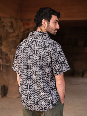 A man with dark hair stands with his back to the camera, wearing a short-sleeved black Shipibo Shirt and green pants, in a sunlit, rustic setting.
