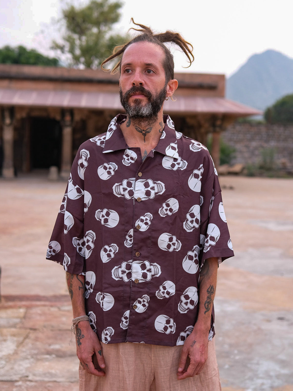 A bearded, tattooed man stands outdoors wearing a stylish Shirova Shirt—a loose short-sleeve shirt in purple with a white skull pattern—and light-colored shorts. Trees, mountains, and an old stone building are visible in the background.