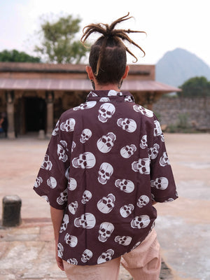 A person with dreadlocks is seen from behind, wearing the loose, stylish Shirova Shirt adorned with a white skull pattern. They stand outdoors near a historical stone building, with hills and trees in the background.